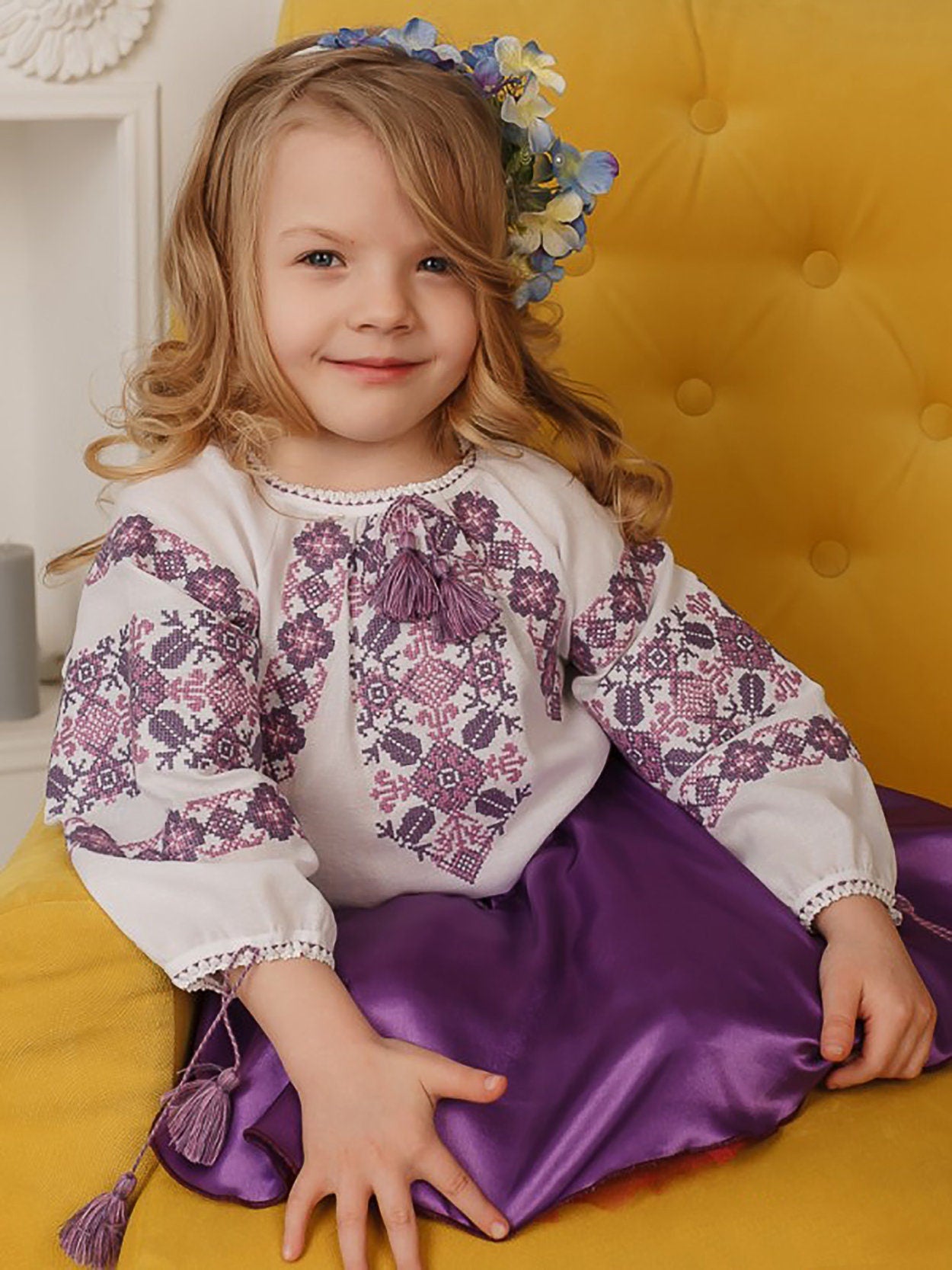 Young girl in traditional embroidered Ukrainian blouse with cross-stitch patterns sitting on yellow chair.
