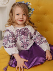 Young girl in traditional embroidered Ukrainian blouse with cross-stitch patterns sitting on yellow chair.