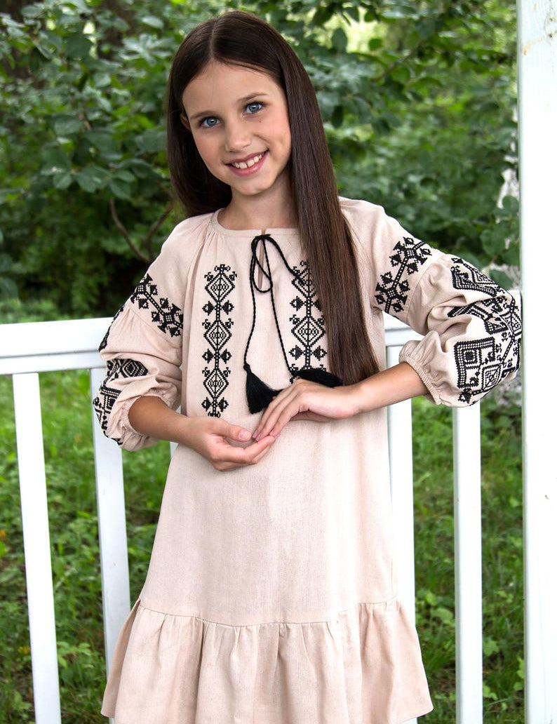 Girl wearing traditional Ukrainian vyshyvanka dress with black embroidered patterns, standing outdoors by a white fence.