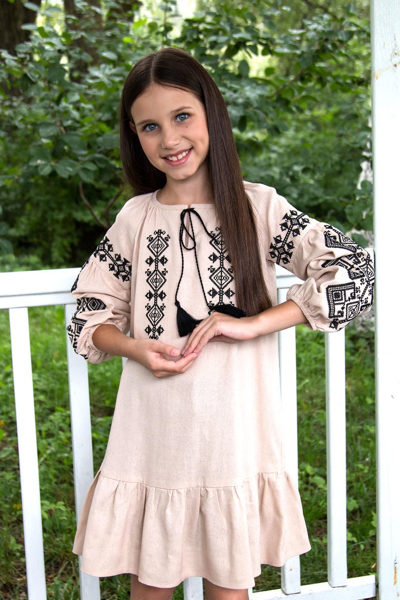 Girl wearing traditional Ukrainian vyshyvanka dress with black embroidered patterns, standing outdoors by a white fence.