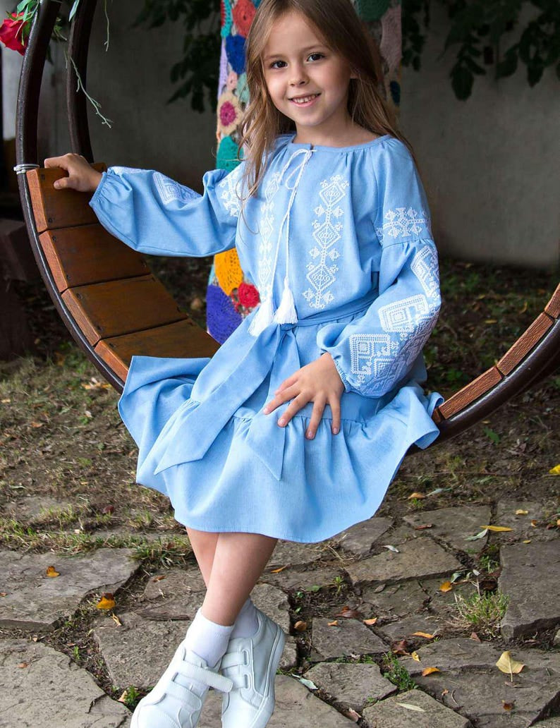 Girl wearing blue embroidered vyshyvanka dress with traditional Ukrainian patterns, sitting outdoors on a wooden swing.