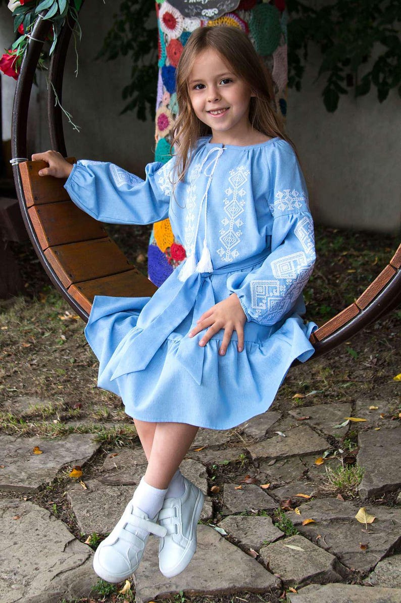 Girl wearing blue embroidered vyshyvanka dress with traditional Ukrainian patterns, sitting outdoors on a wooden swing.