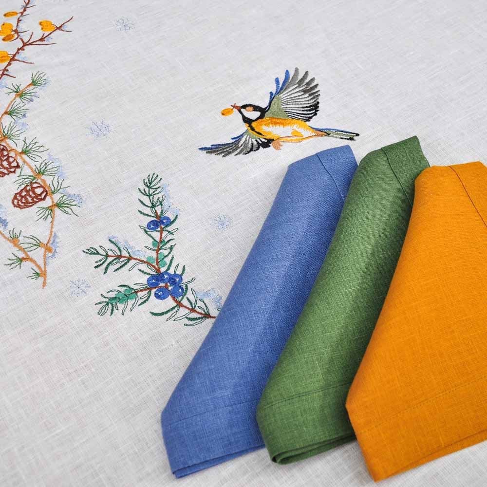 Embroidered linen tablecloth with bird and pine motifs, featuring blue, green, and yellow napkins.