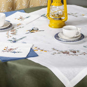 Embroidered linen tablecloth set with floral design, featuring a 90x90 cm cloth and napkins, styled with teacups and a yellow lantern.