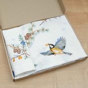 Embroidered linen tablecloth and napkin set with a bird and pine cone design, displayed in an open box on a wooden surface.