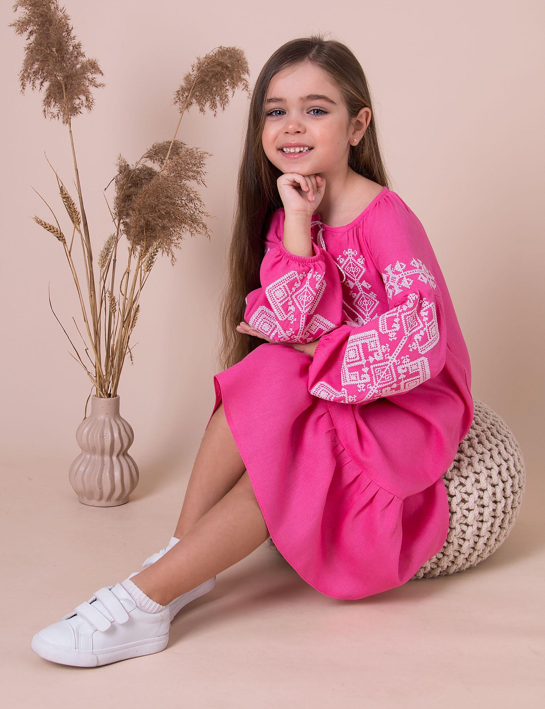 Girl wearing pink embroidered vyshyvanka dress with traditional Ukrainian patterns, sitting in a stylish setting.
