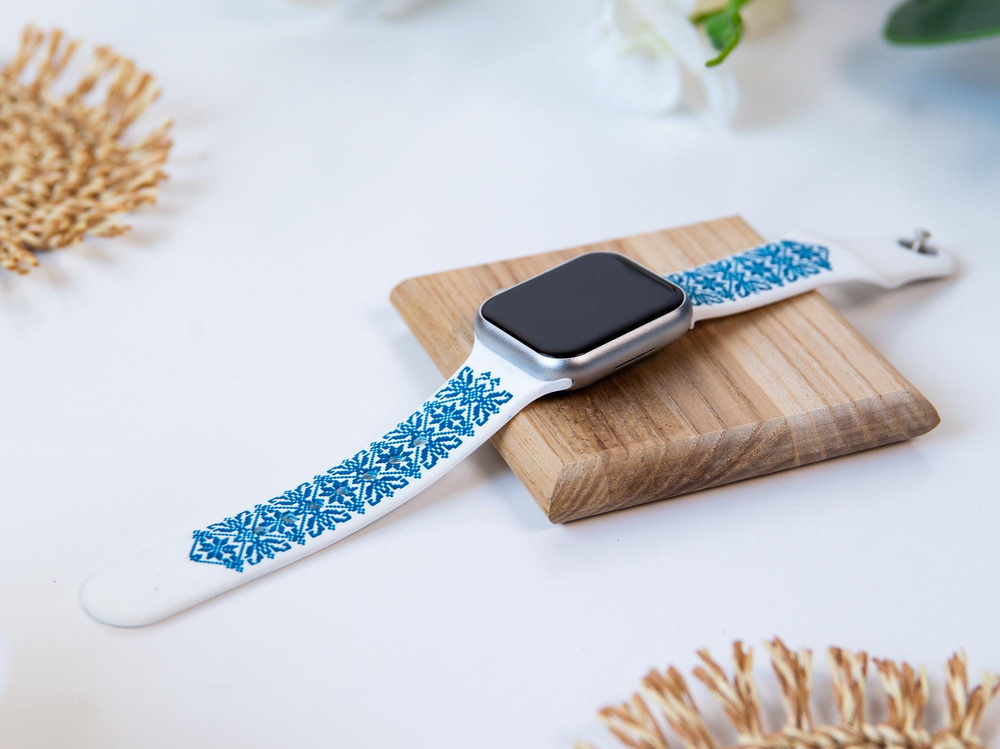 Elegant white and blue Apple Watch band with Ukrainian embroidery design, displayed on a wooden platform.