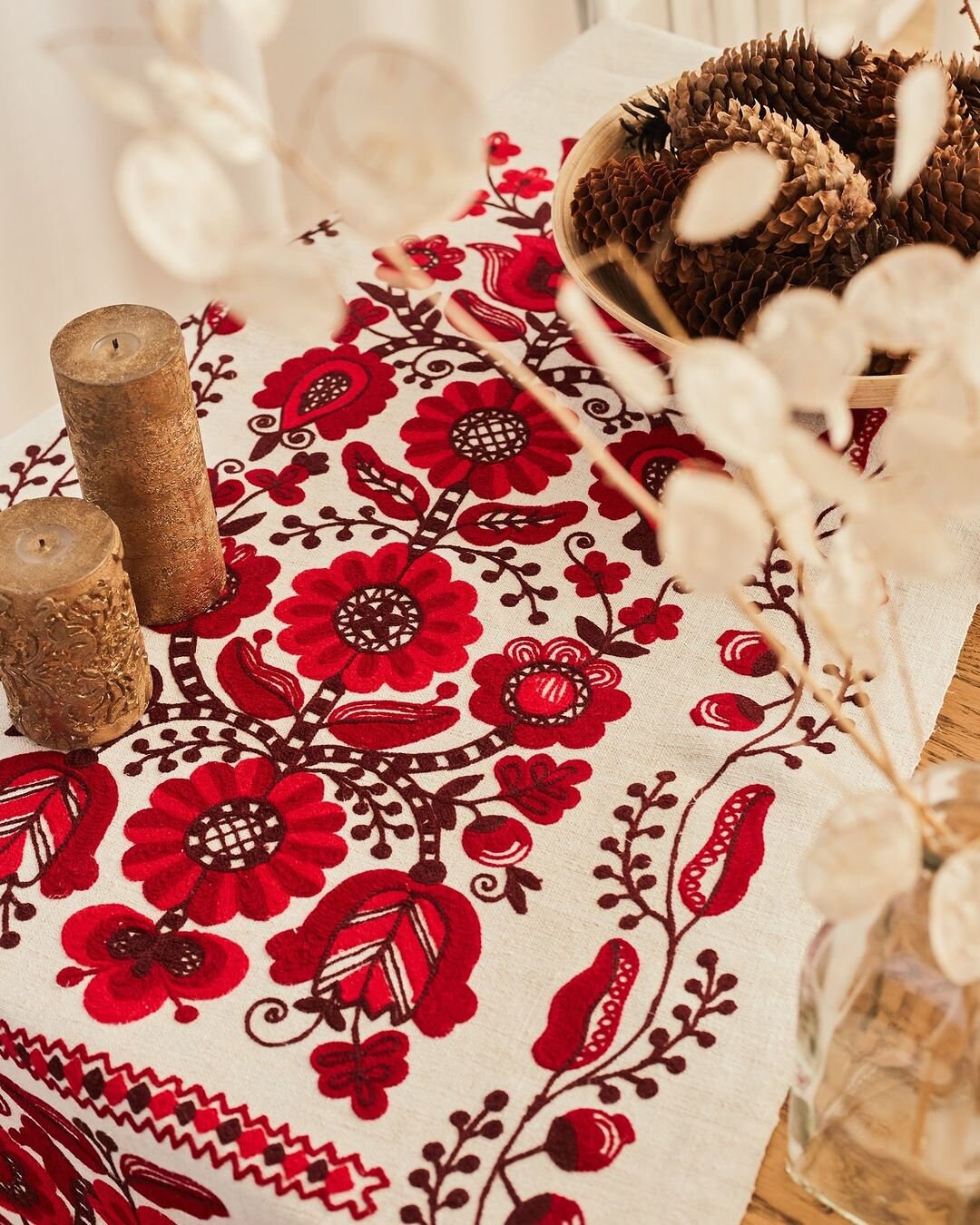 Ukrainian embroidered rushnyk on table with red cross-stitched floral motifs, pinecones, candles, and dried floral decor.
