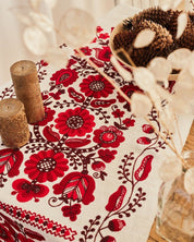 Ukrainian embroidered rushnyk on table with red cross-stitched floral motifs, pinecones, candles, and dried floral decor.