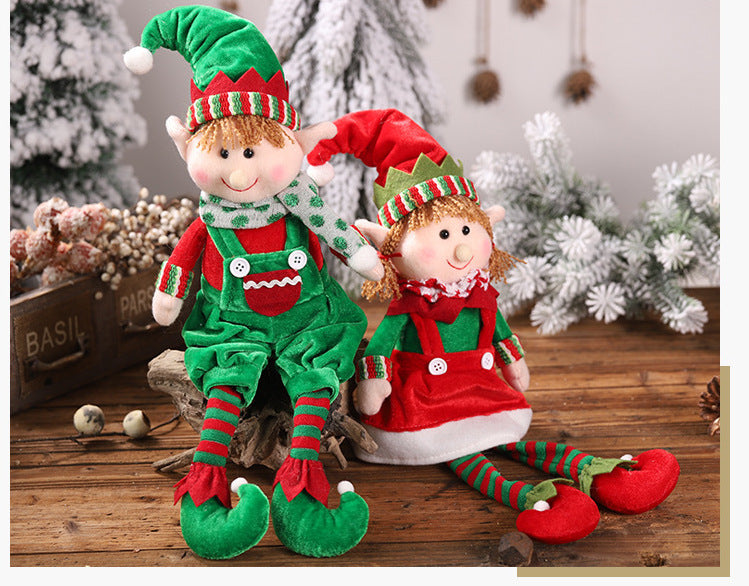 Christmas hanging legs sitting dolls, male and female models, on holiday-decorated table with snow-covered trees in background.