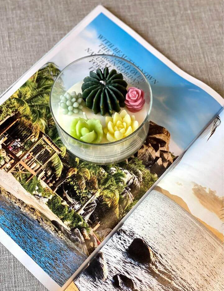 Garden Escape terrarium candle with floral wax designs on a magazine showcasing a tropical beach scene.