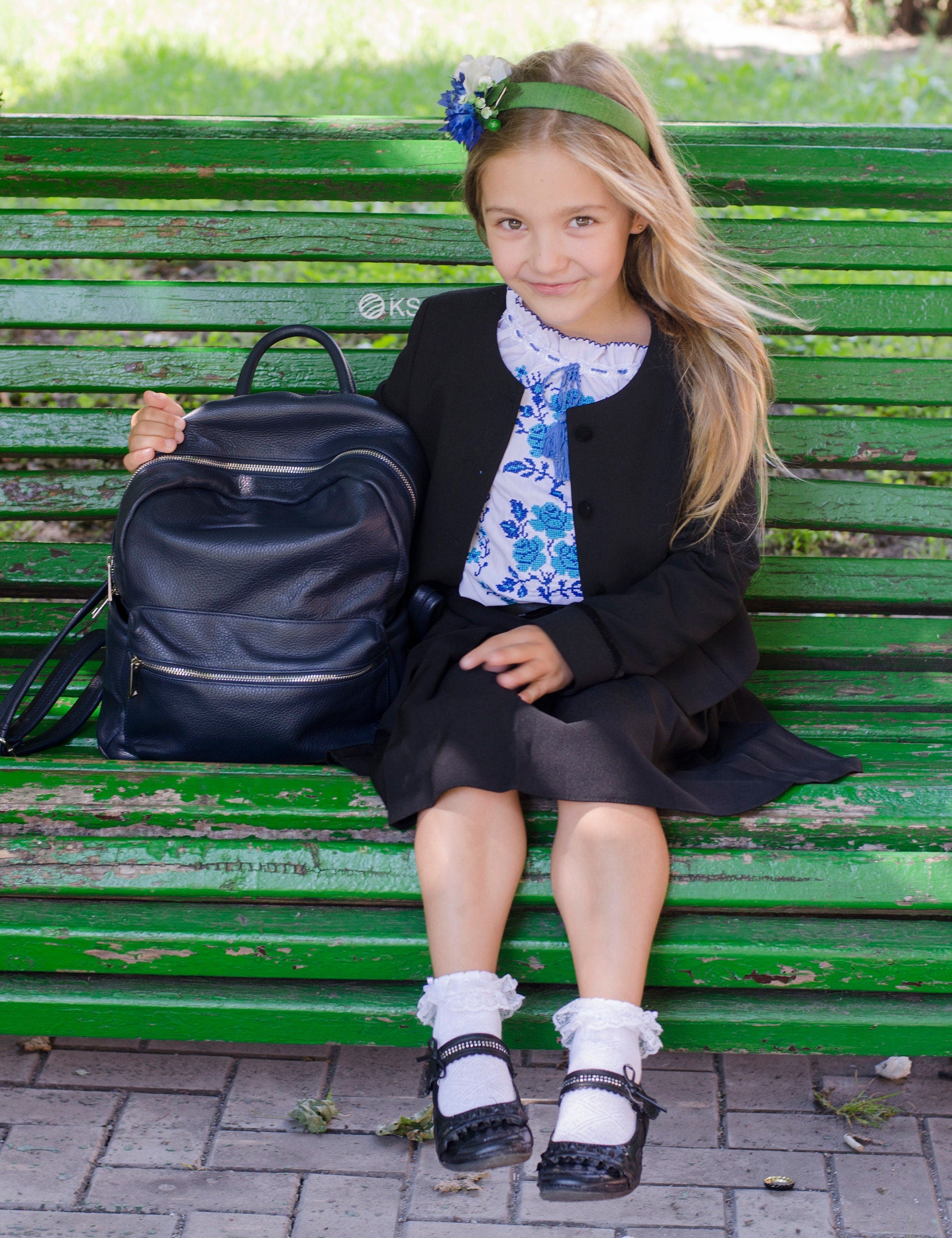 Girl in school uniform sitting on a green bench with a backpack, wearing a floral blouse and black jacket, outdoors in a park.