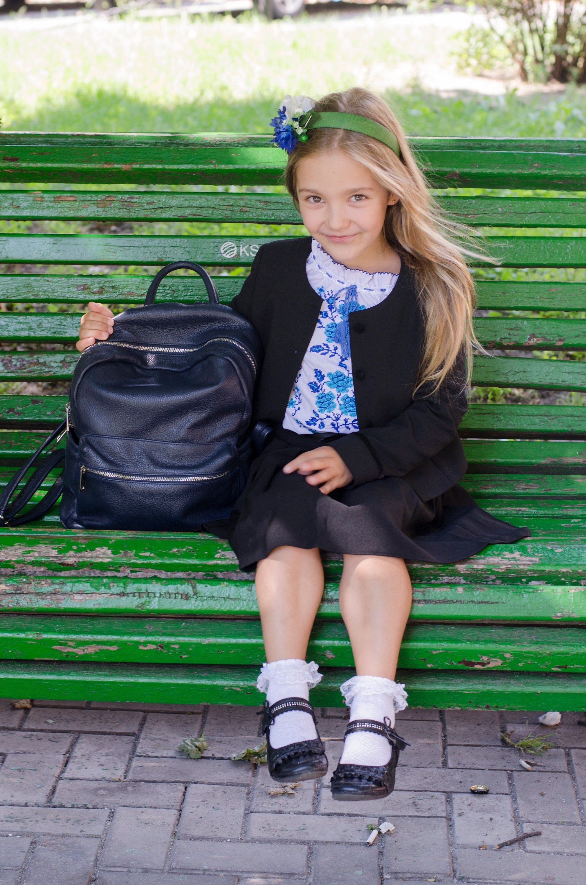 Girl in school uniform sitting on a green bench with a backpack, wearing a floral blouse and black jacket, outdoors in a park.