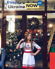 Girl wearing a white embroidered Vyshyvanka dress with floral cross-stitch, standing in front of a festive shop window.