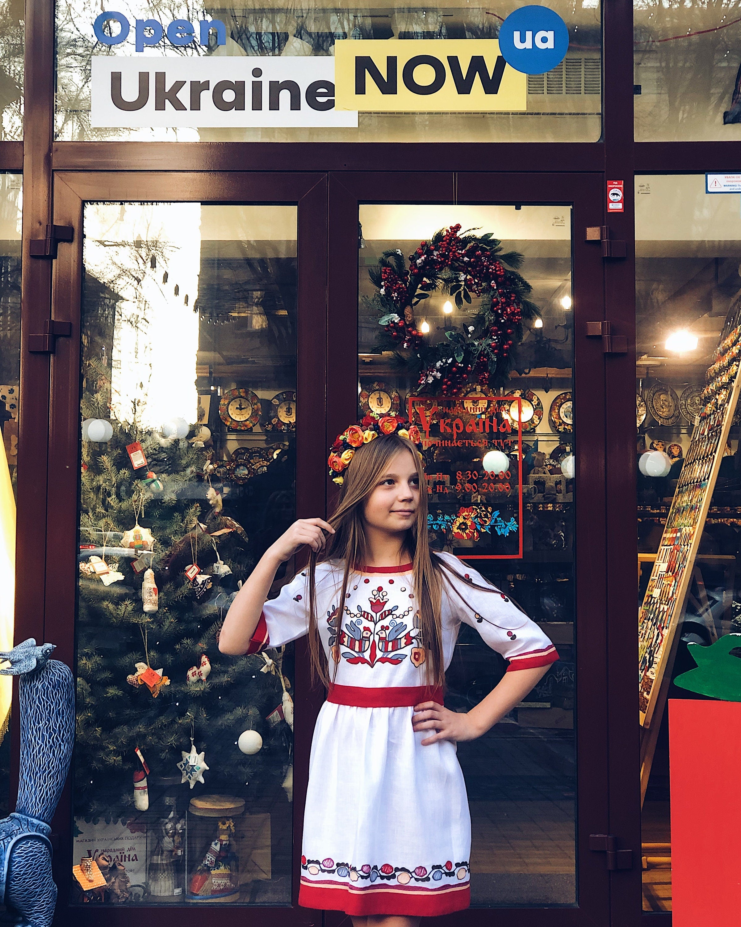 Girl wearing a white embroidered Vyshyvanka dress with floral cross-stitch, standing in front of a festive shop window.