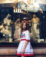 Girl wearing white embroidered dress with floral cross-stitch, standing beside nativity scene, showcasing Ukrainian Vyshyvanka tradition.