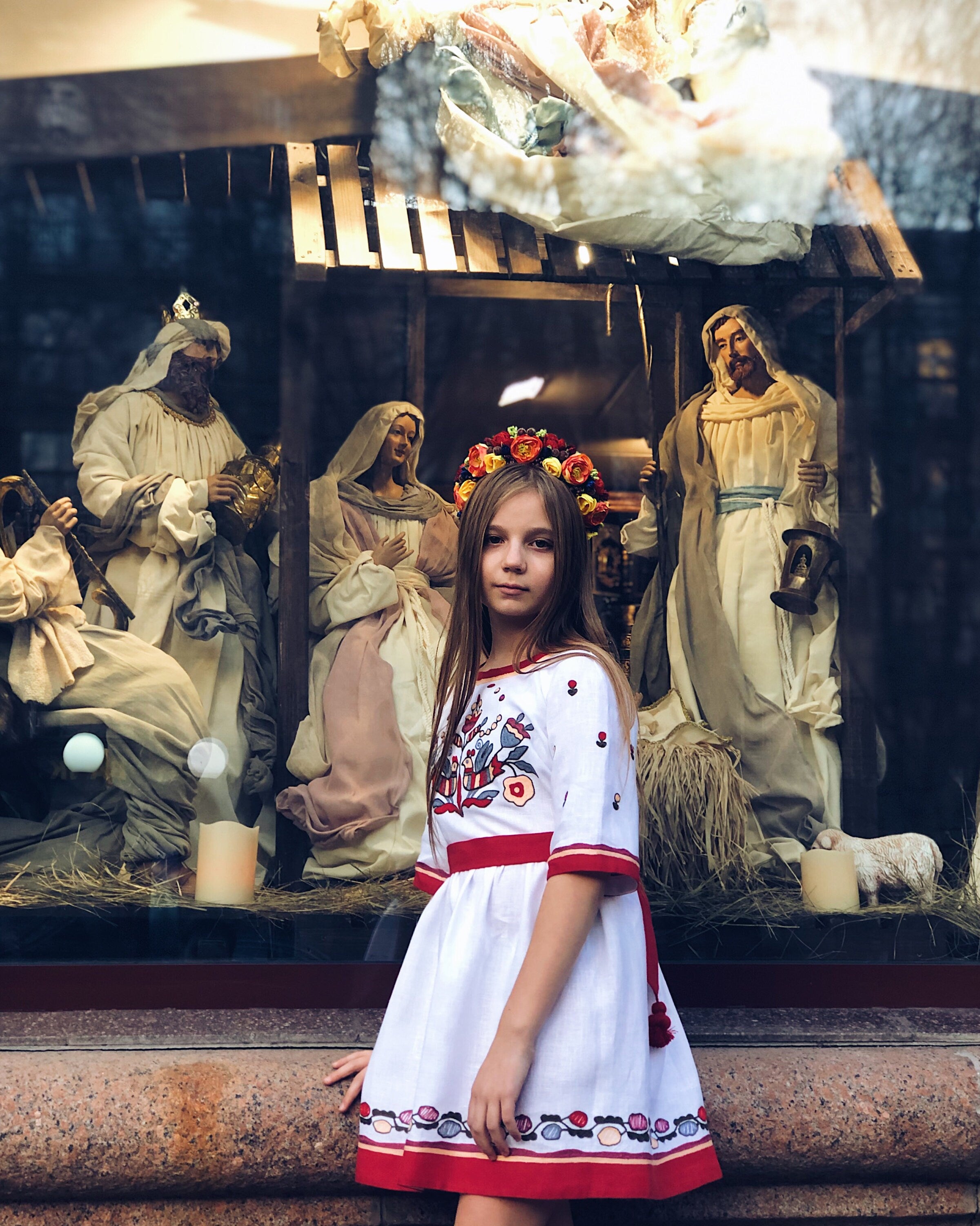 Girl wearing white embroidered dress with floral cross-stitch, standing beside nativity scene, showcasing Ukrainian Vyshyvanka tradition.