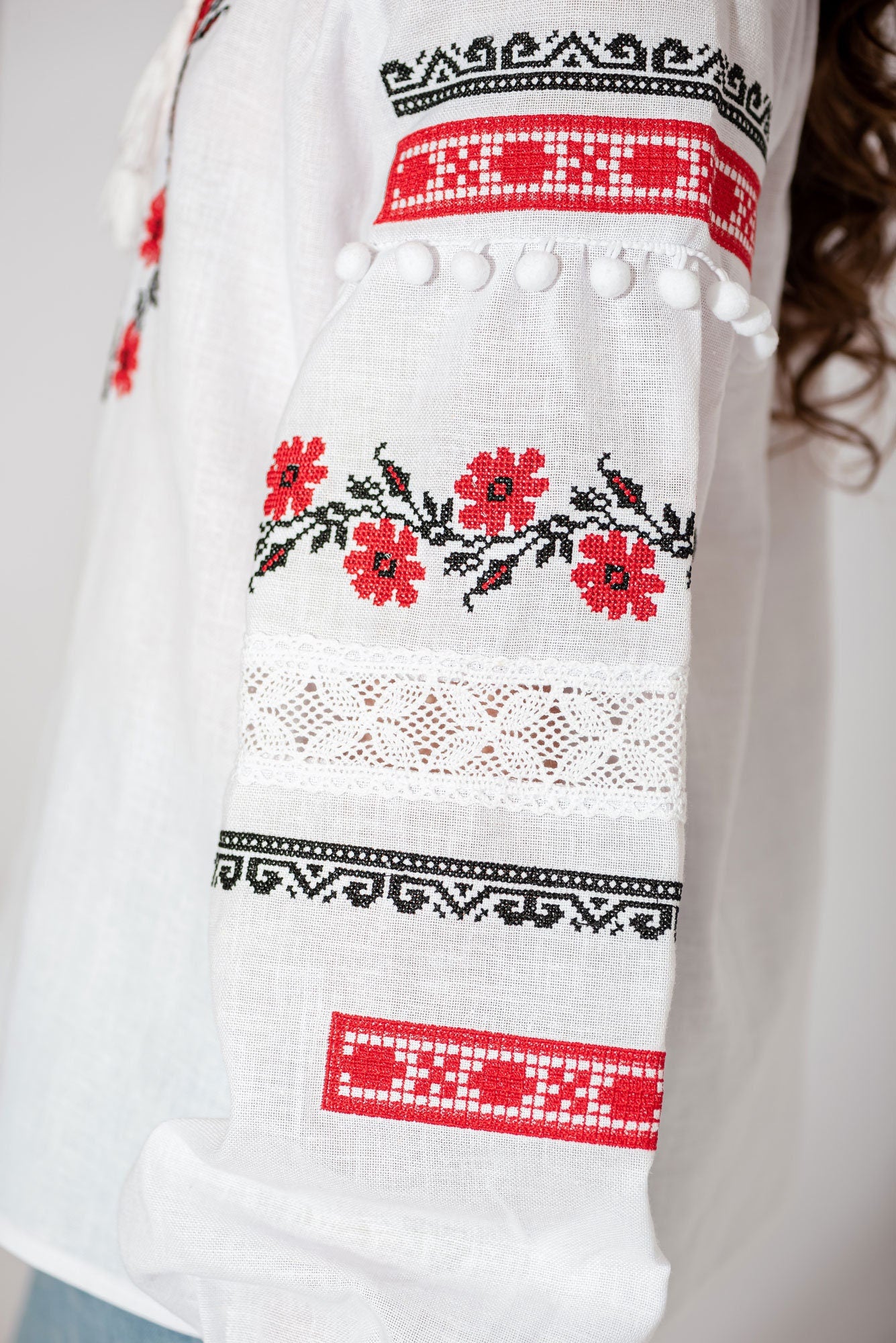 Close-up of girls' white Vyshyvanka blouse sleeve with red and black floral embroidery, lace accents, and pom-poms.