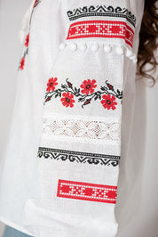 Close-up of girls' white Vyshyvanka blouse sleeve with red and black floral embroidery, lace accents, and pom-poms.