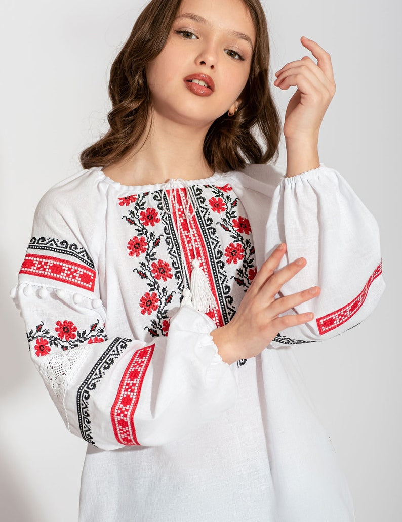 Girl wearing a white Ukrainian vyshyvanka blouse with red and black floral embroidery, lace accents, and pom-pom sleeves.
