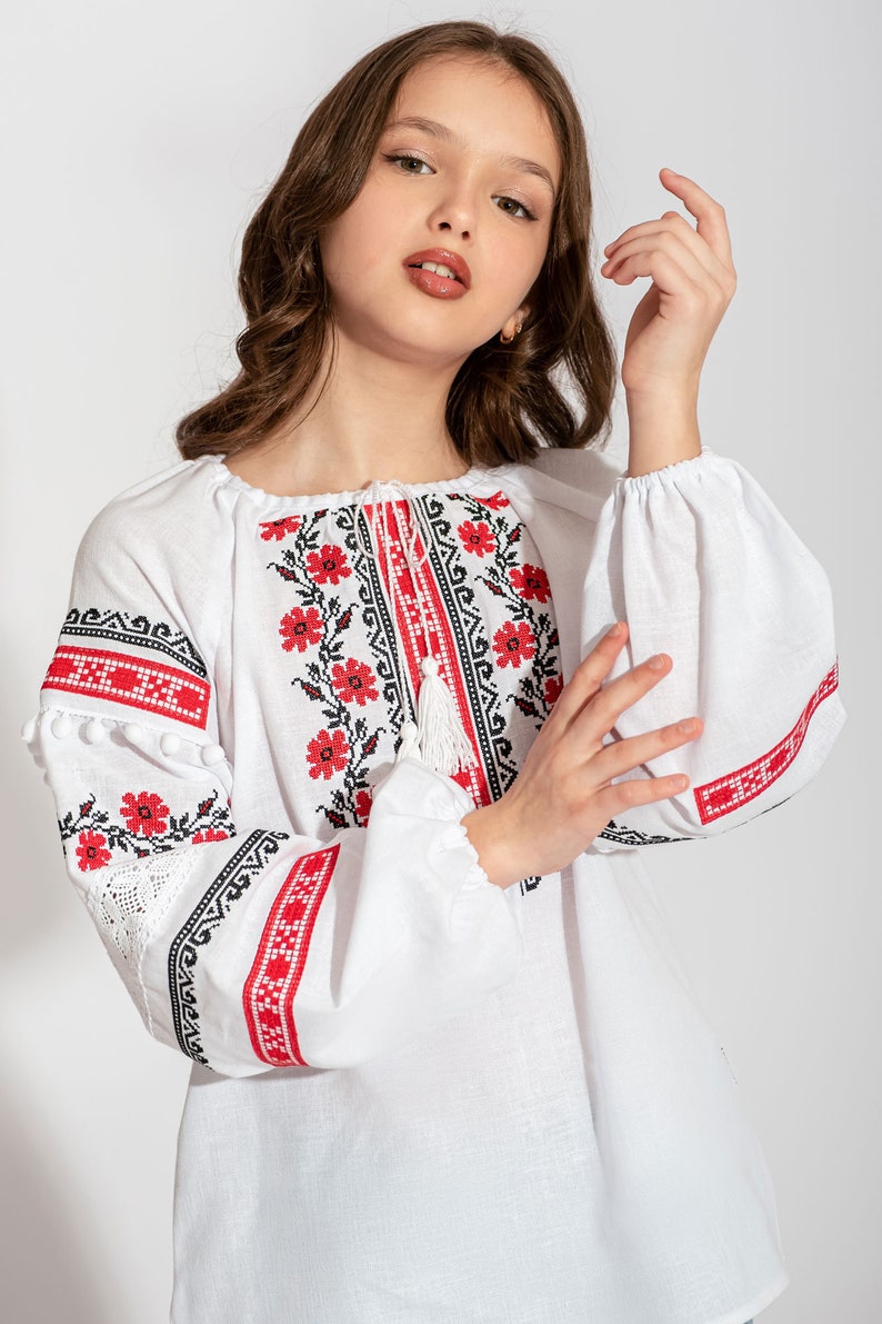 Girl wearing a white Ukrainian vyshyvanka blouse with red and black floral embroidery, lace accents, and pom-pom sleeves.
