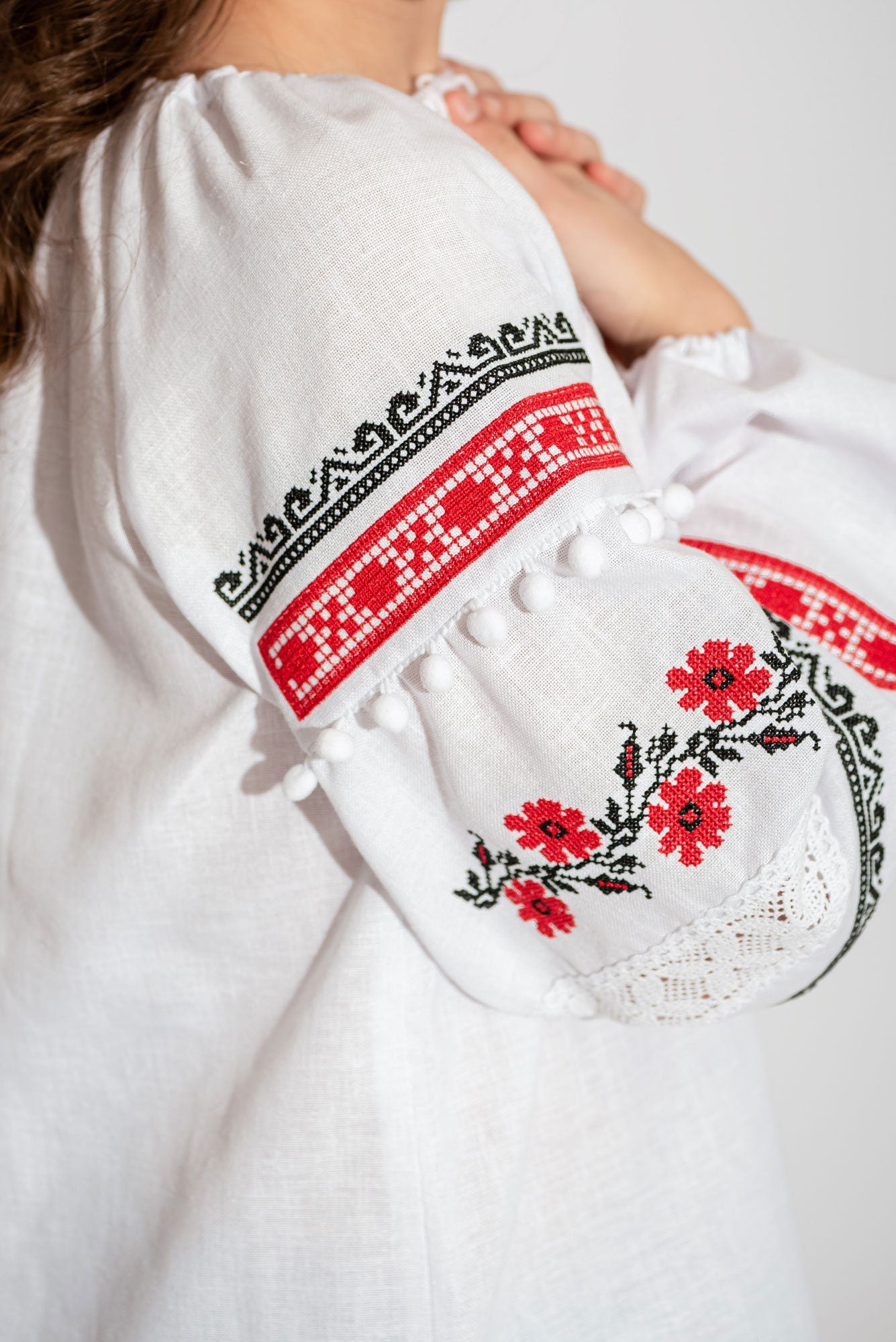 Girls’ embroidered vyshyvanka blouse with red and black floral pattern on white linen cotton, featuring lace and pom-poms.