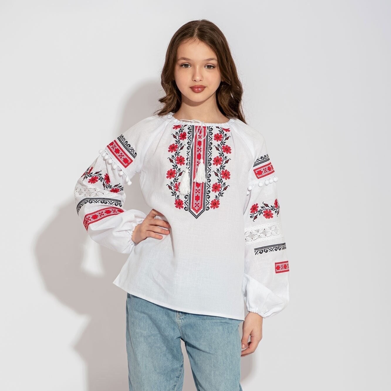 Girls' white vyshyvanka blouse with red and black floral embroidery, crafted from cotton linen, featuring lace and pom-pom details.