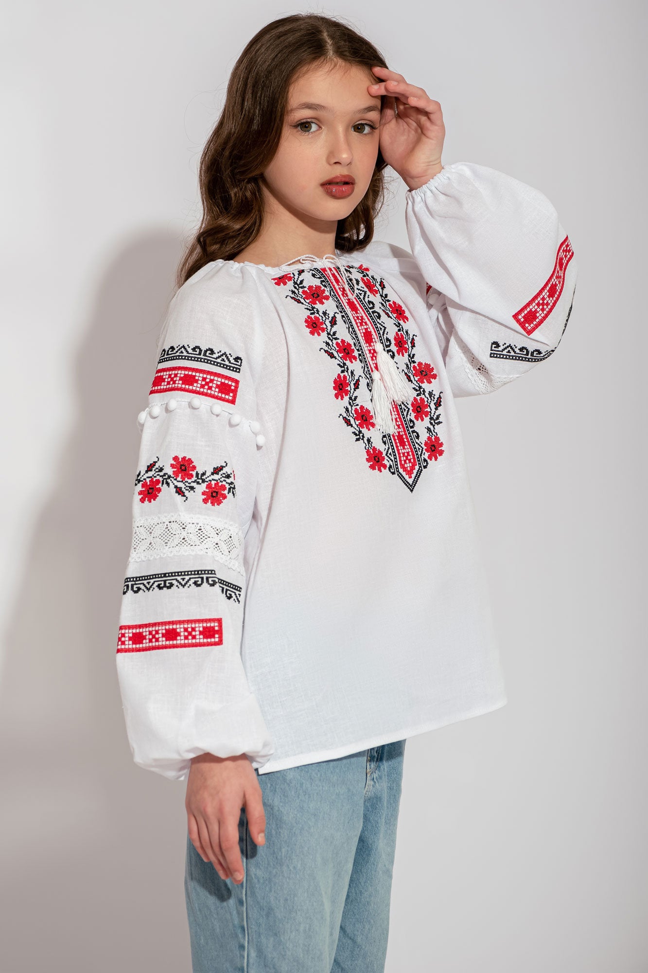 Girls' white embroidered Ukrainian vyshyvanka blouse with red and black floral patterns, featuring lace and sleeve pom-poms.
