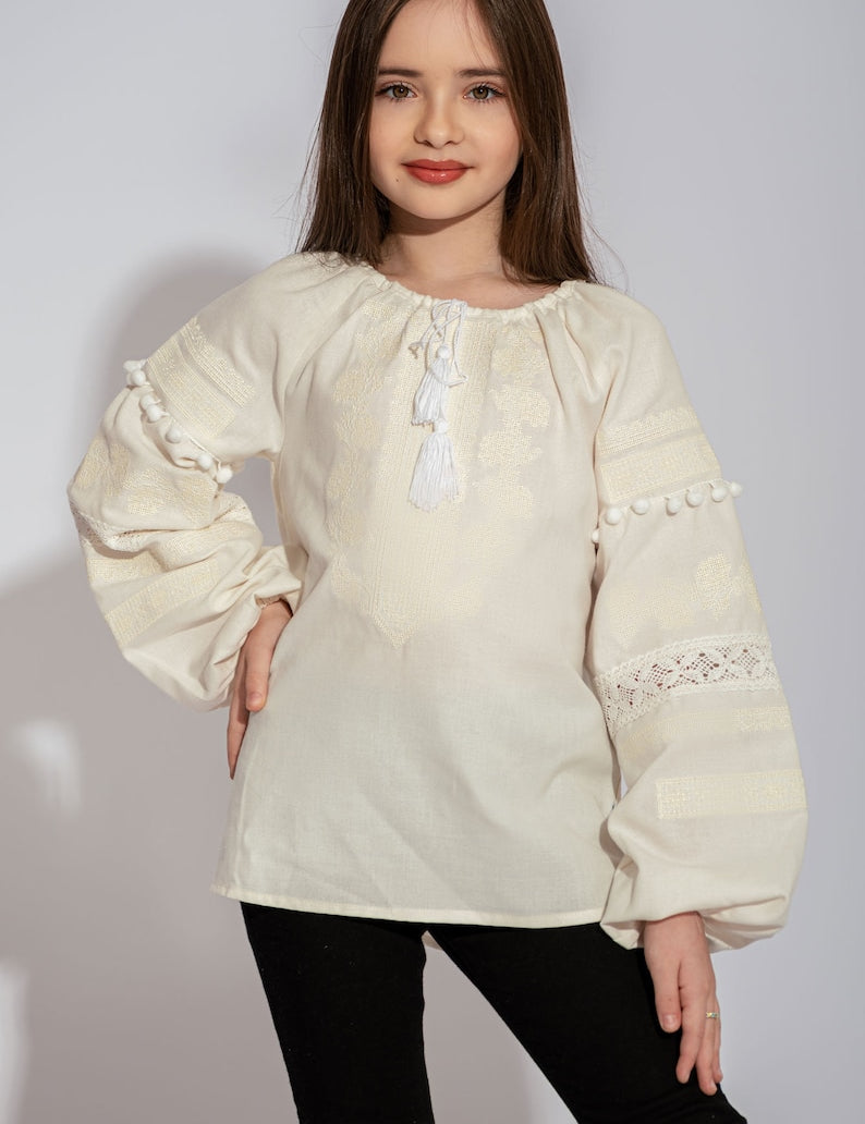Girl wearing a white Vyshyvanka blouse with red and black floral embroidery, lace accents, and pom-poms. Traditional Ukrainian shirt.