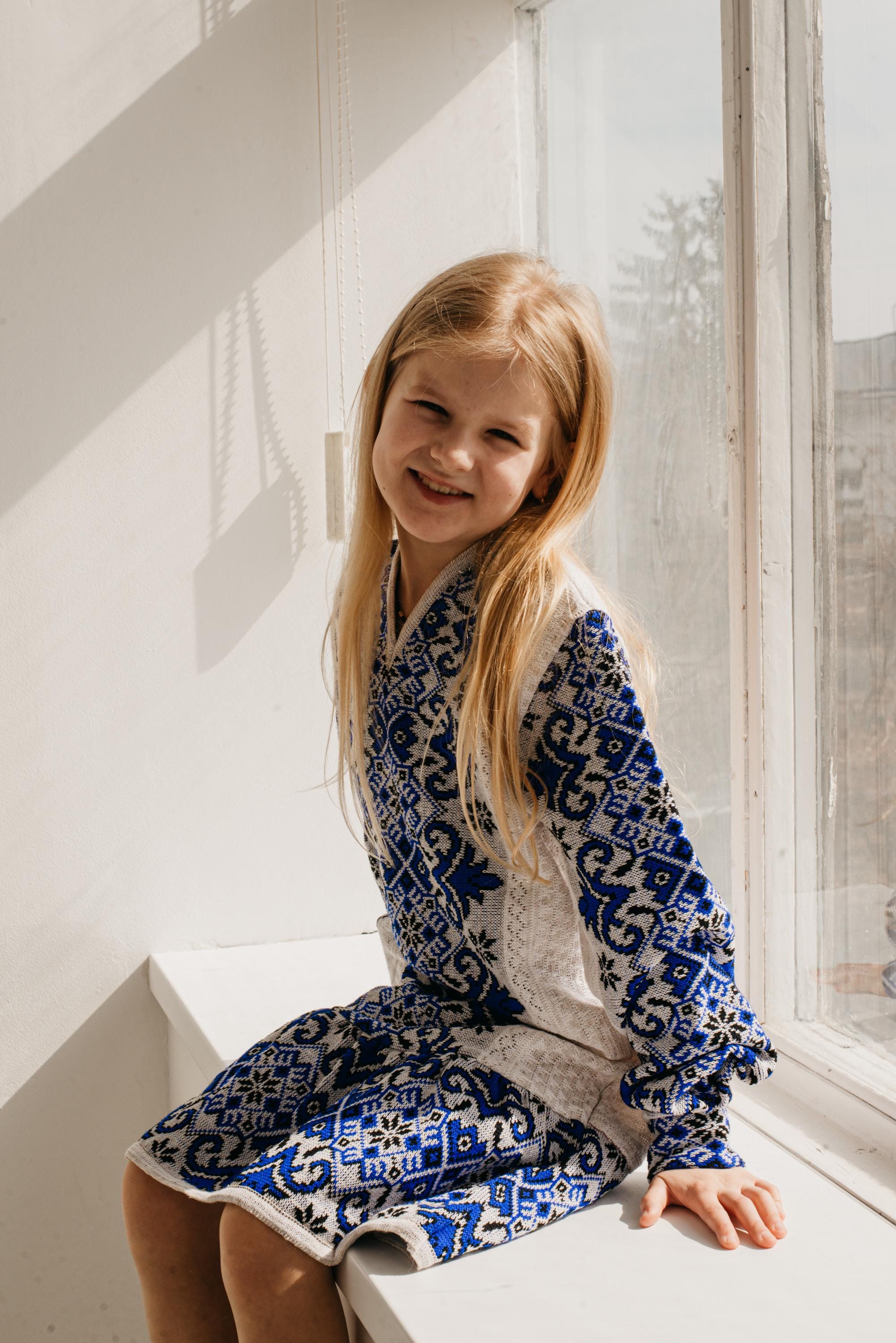 Young girl in traditional Ukrainian embroidered knitted dress sitting by window, smiling.