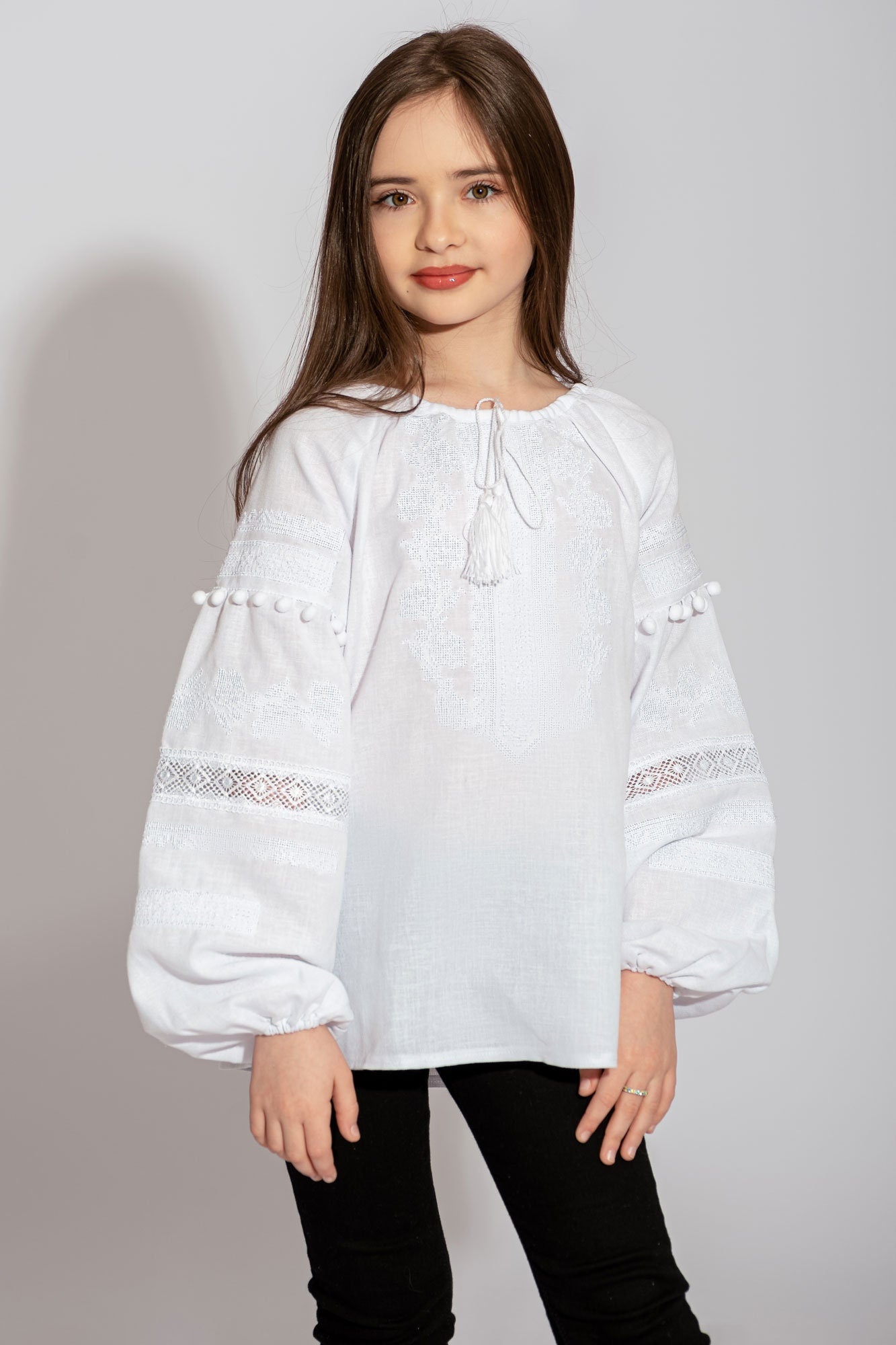 Girl wearing white embroidered vyshyvanka blouse with red and black floral pattern, lace accents, and sleeve pom-poms.