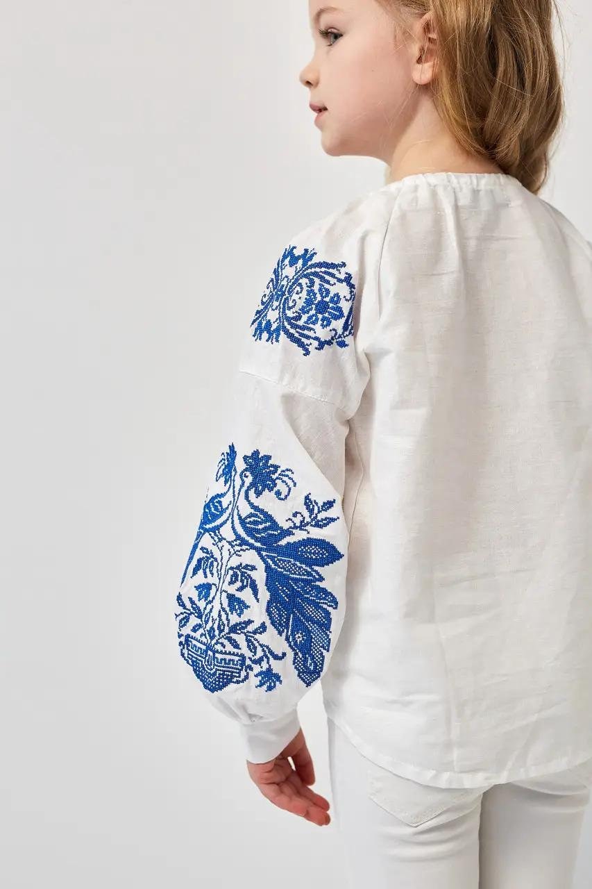 Girl wearing white Ukrainian embroidered shirt with blue phoenix motifs, 100% cotton, symbolizing heritage and strength.