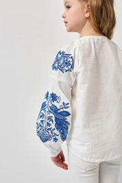 Girl wearing white Ukrainian embroidered shirt with blue phoenix motifs, 100% cotton, symbolizing heritage and strength.