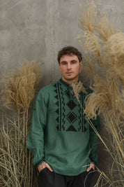 Man wearing green embroidered Ukrainian linen shirt, traditional vyshyvanka, standing by pampas grass against concrete wall.