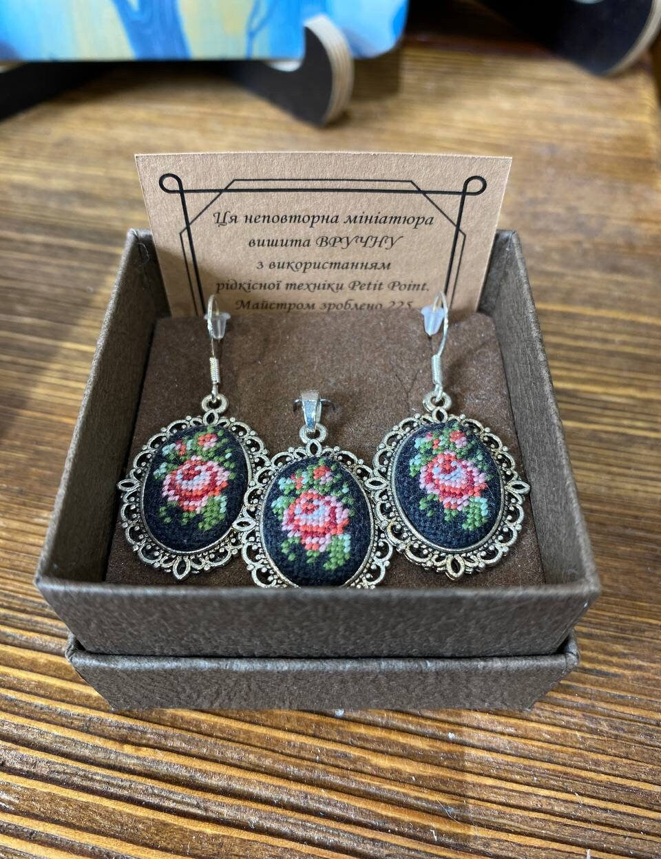 Hand-embroidered jewelry set with floral Petit Point silk pendant and earrings displayed in a box, showcasing intricate craftsmanship.