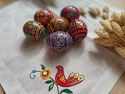 Set of 6 hand-painted Ukrainian pysanky wooden eggs with intricate folk designs, displayed on embroidered cloth with a bird motif.