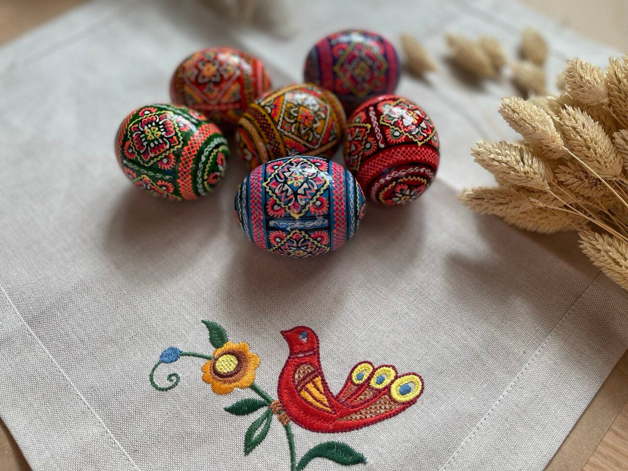 Set of 6 hand-painted Ukrainian pysanky wooden eggs with intricate folk designs, displayed on embroidered cloth with a bird motif.