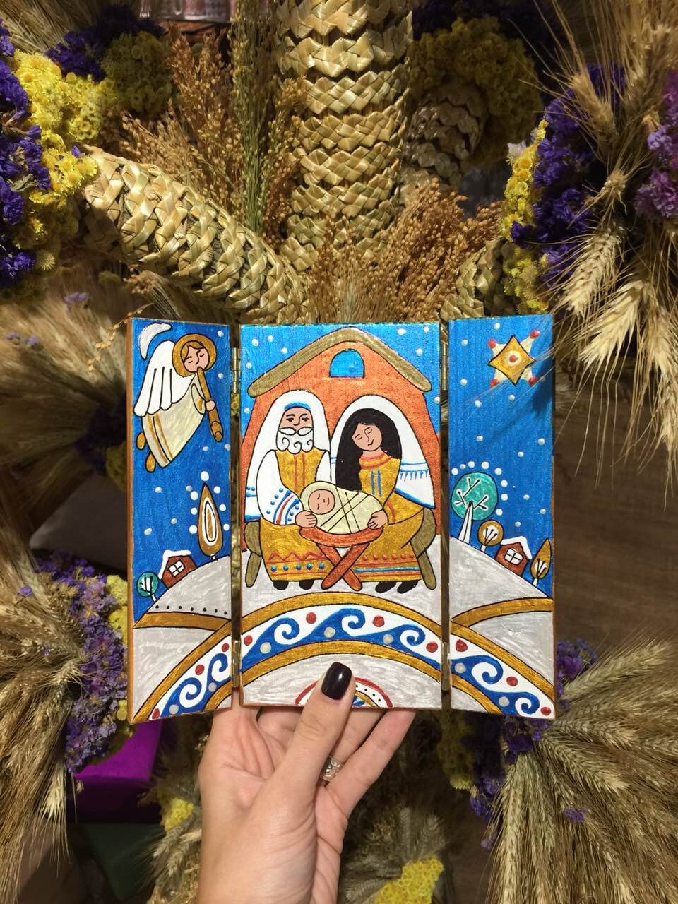 Hand holding a hand-painted wooden icon depicting a religious scene with vibrant colors, surrounded by dried flowers.