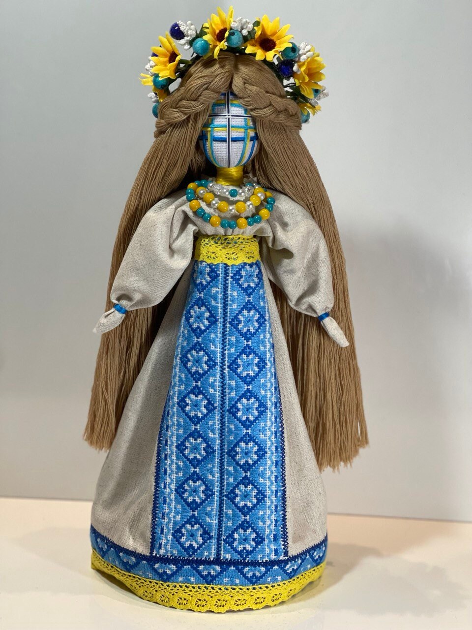 Handmade Ukrainian Motanka doll with blue embroidered dress, floral headpiece on wooden stand, symbolizing heritage and well-being.
