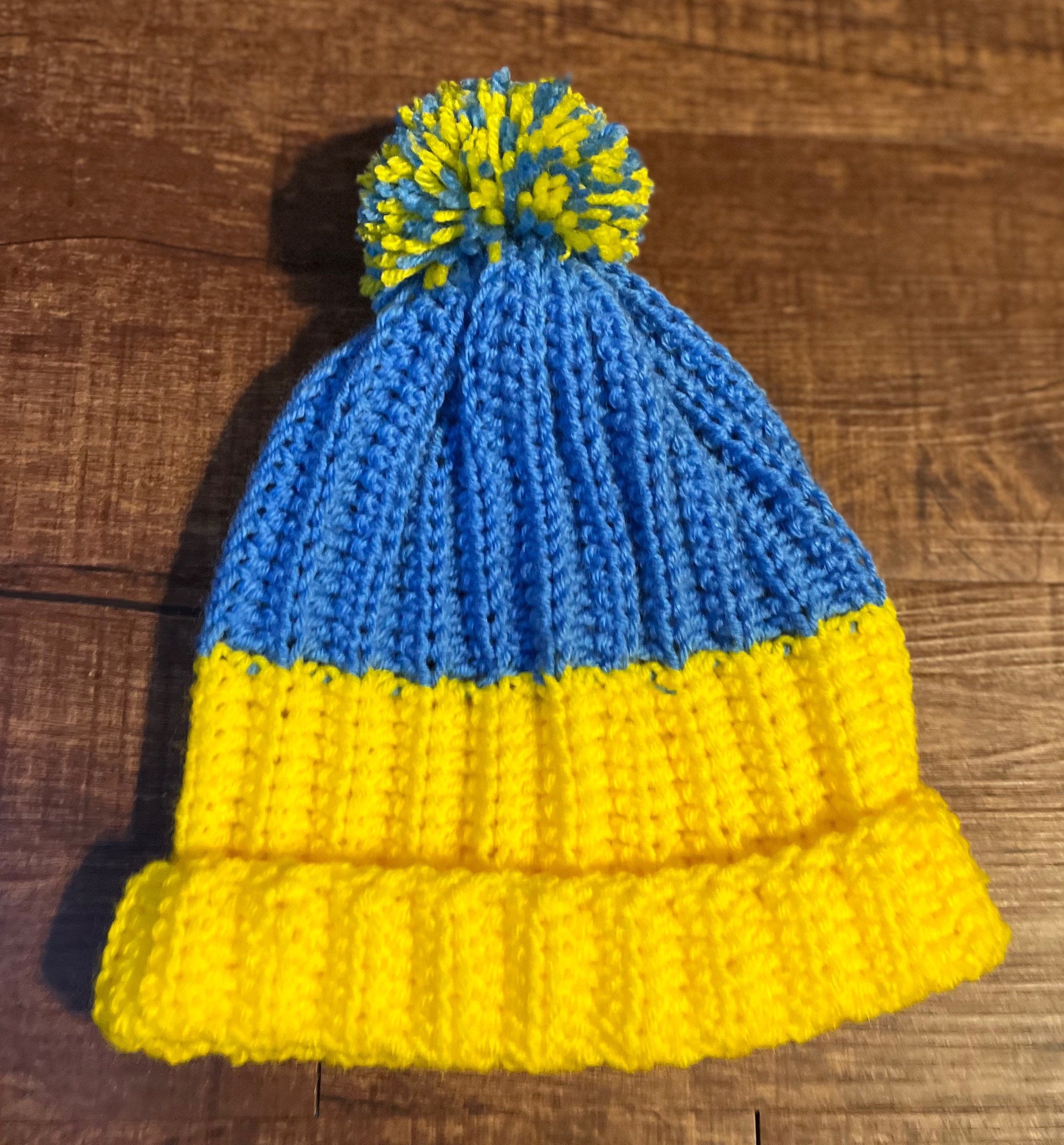 Handmade blue and yellow crochet hat, Ukraine winter support accessory featuring pom-pom and flag colors on wooden background.