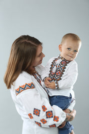 Mother holding child wearing traditional Ukrainian embroidered shirts with vibrant cross-stitch patterns.