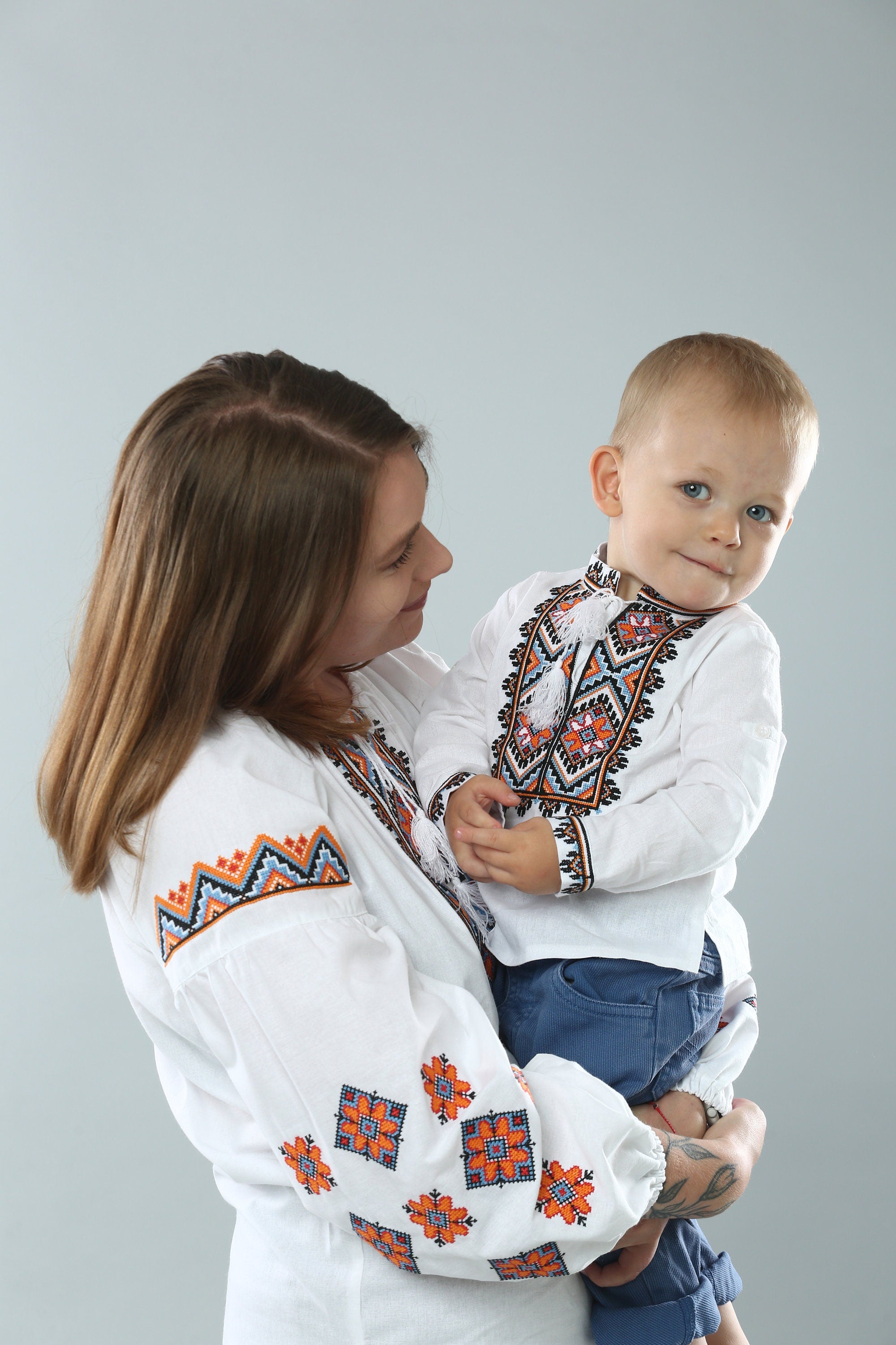 Mother holding child wearing traditional Ukrainian embroidered shirts with vibrant cross-stitch patterns.