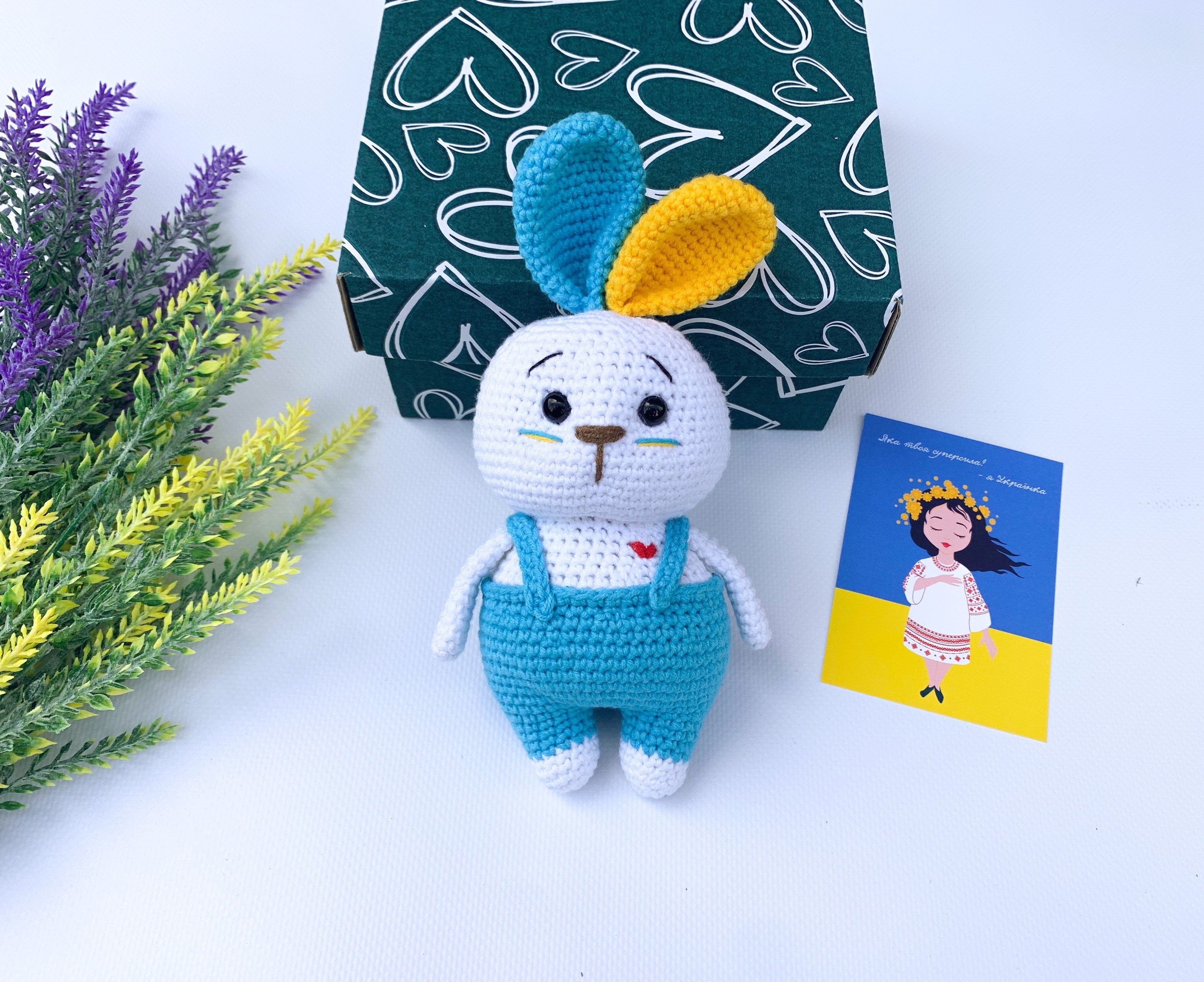 Handmade Ukrainian crochet bunny toy with colorful ears and blue overalls, perfect Easter or baby shower gift, next to a gift box.