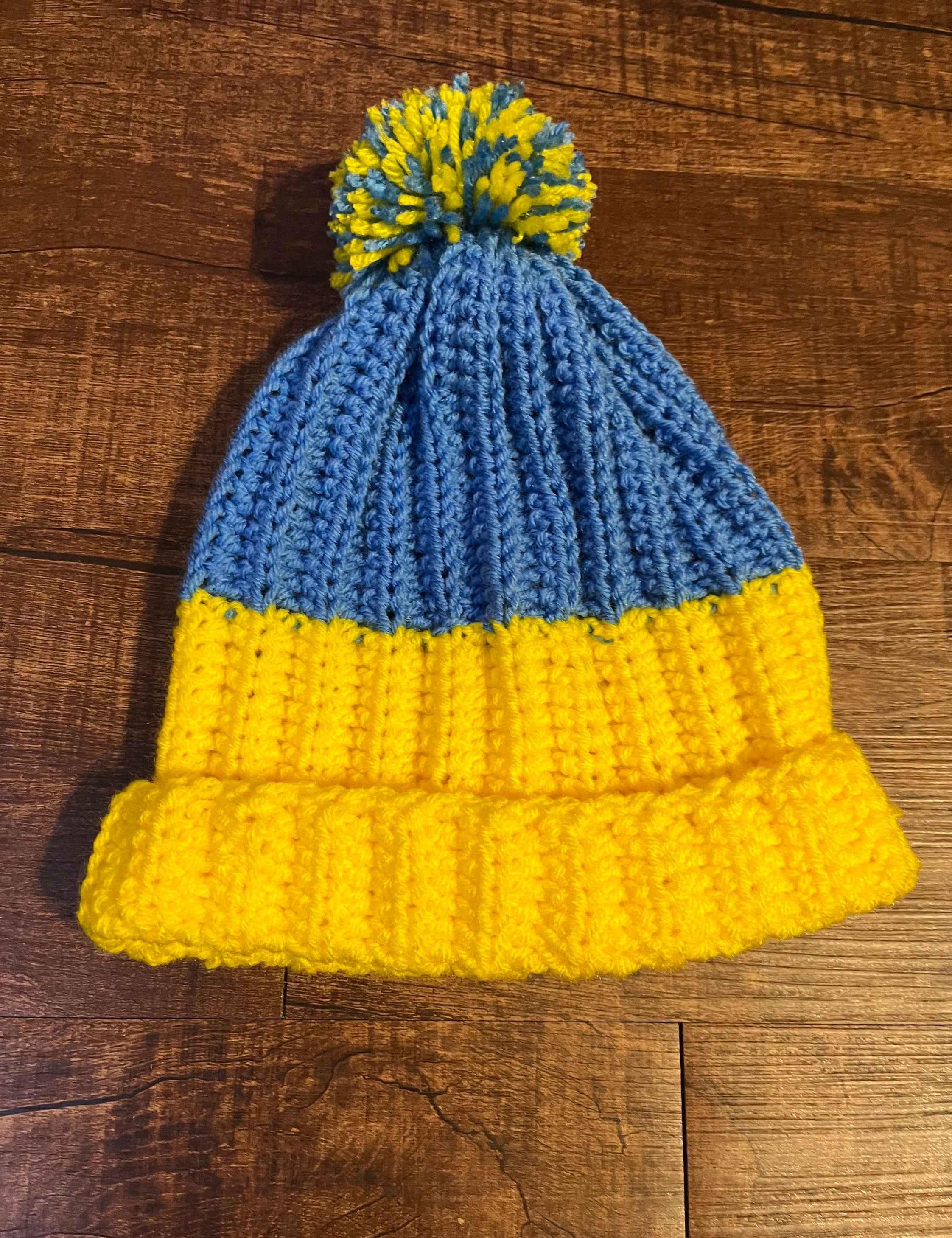 Handmade blue and yellow crochet hat with pom-pom, symbolizing support for Ukraine, on a wooden background.