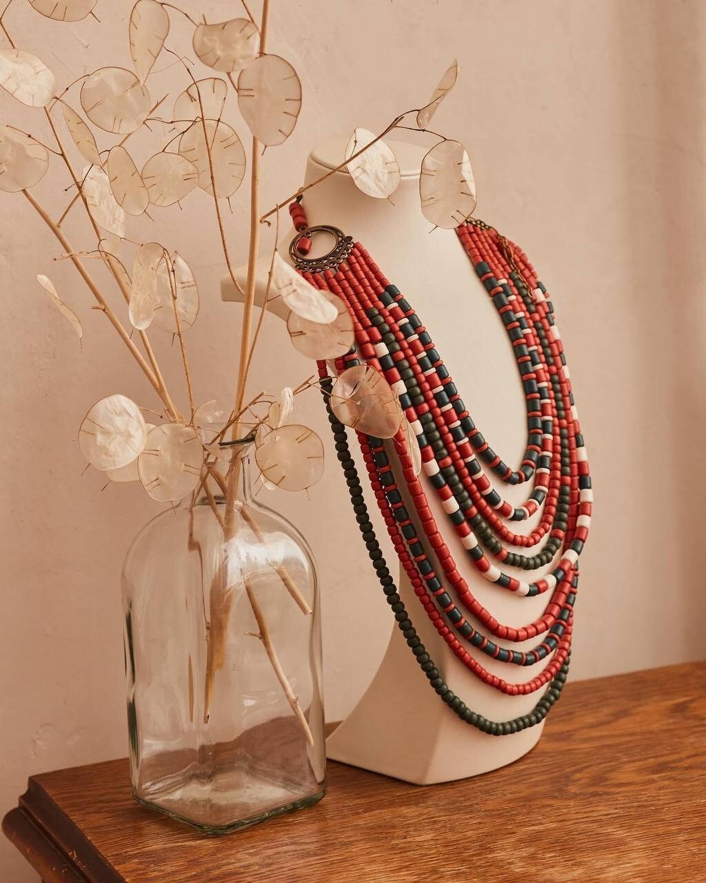 Decorative layered beaded necklaces in red and black on a stand beside a vase with dried plant stems.