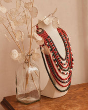 Decorative layered beaded necklaces in red and black on a stand beside a vase with dried plant stems.