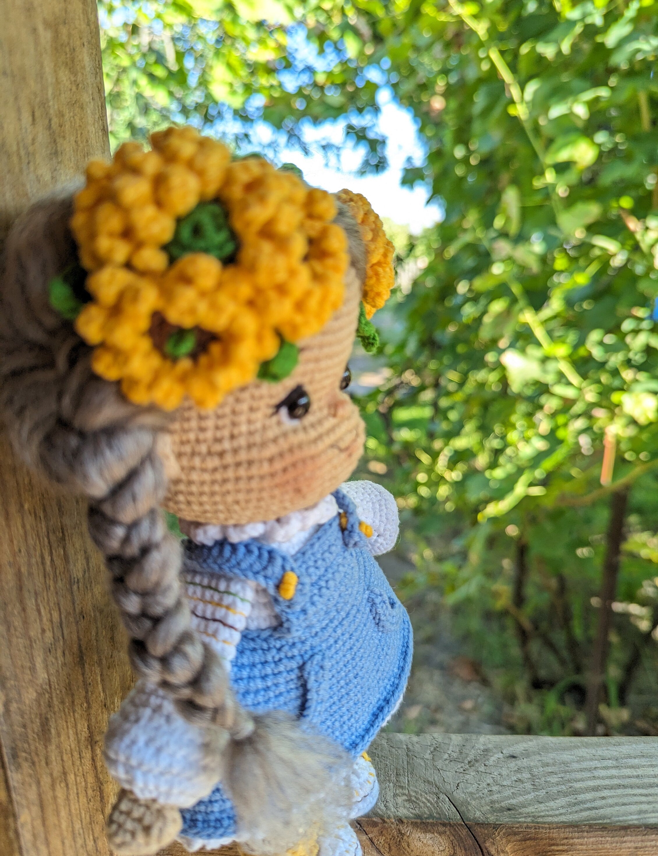 Crochet Ukrainian doll in blue dress with yellow flower crown, folk-style soft toy, against a green leafy background.