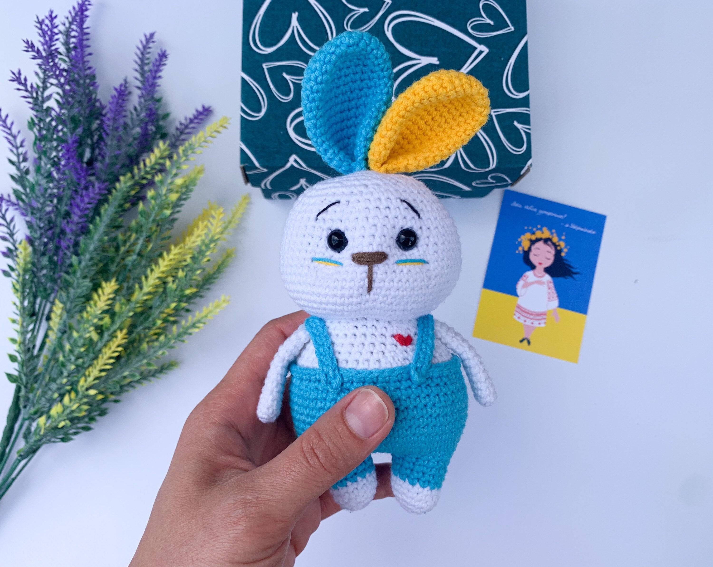 Handmade Ukrainian crochet bunny toy with blue overalls, a multicolored ear, and a heart detail, perfect gift for baby showers or Easter.