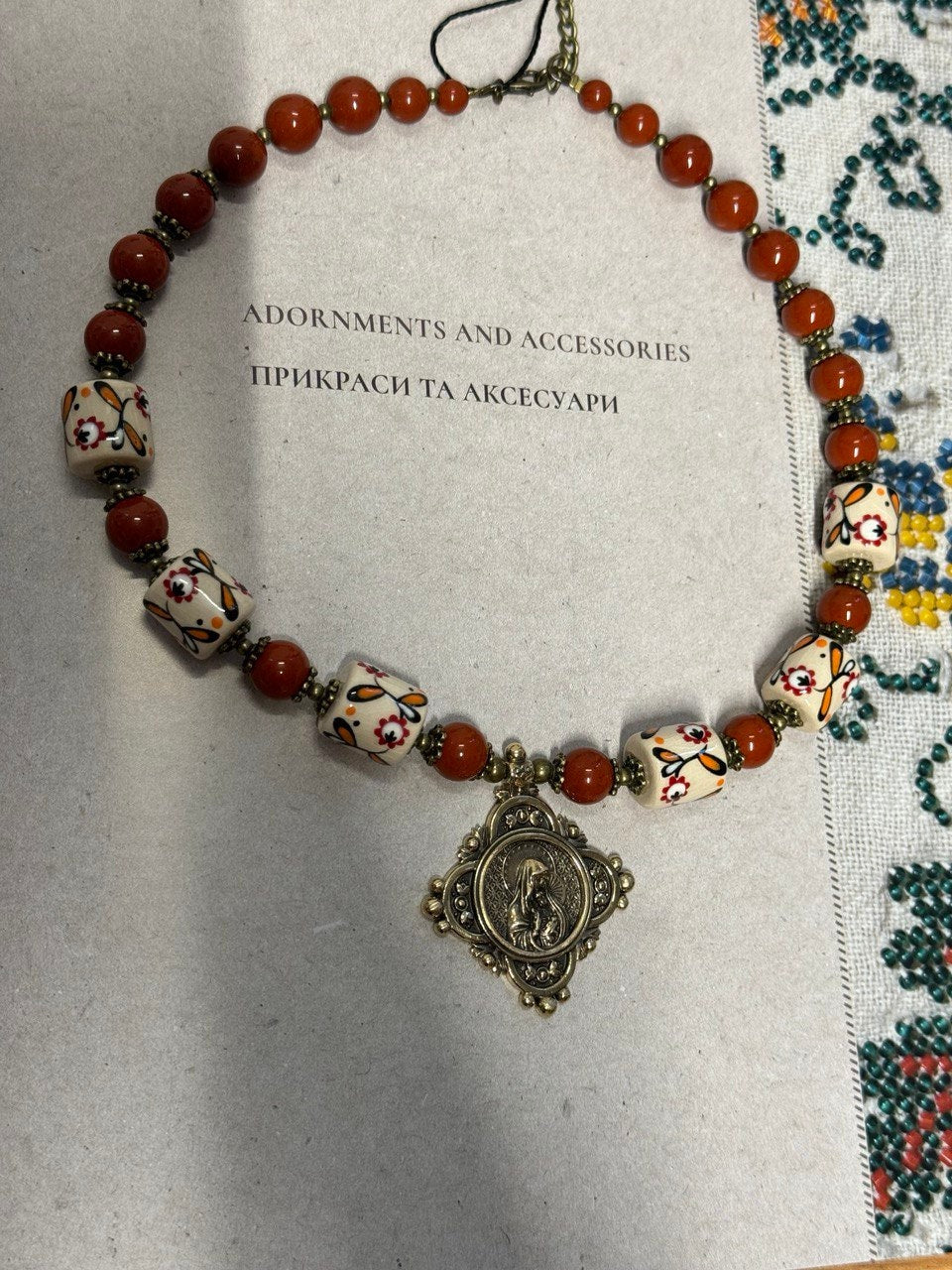 Handmade Ukrainian necklace with white clay, jasper beads, and brass medallion on traditional embroidered fabric background.