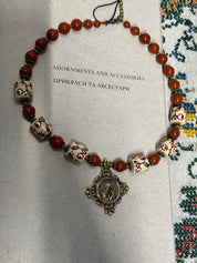 Ukrainian handmade necklace with white clay and red jasper beads, featuring brass pendant on traditional fabric background.